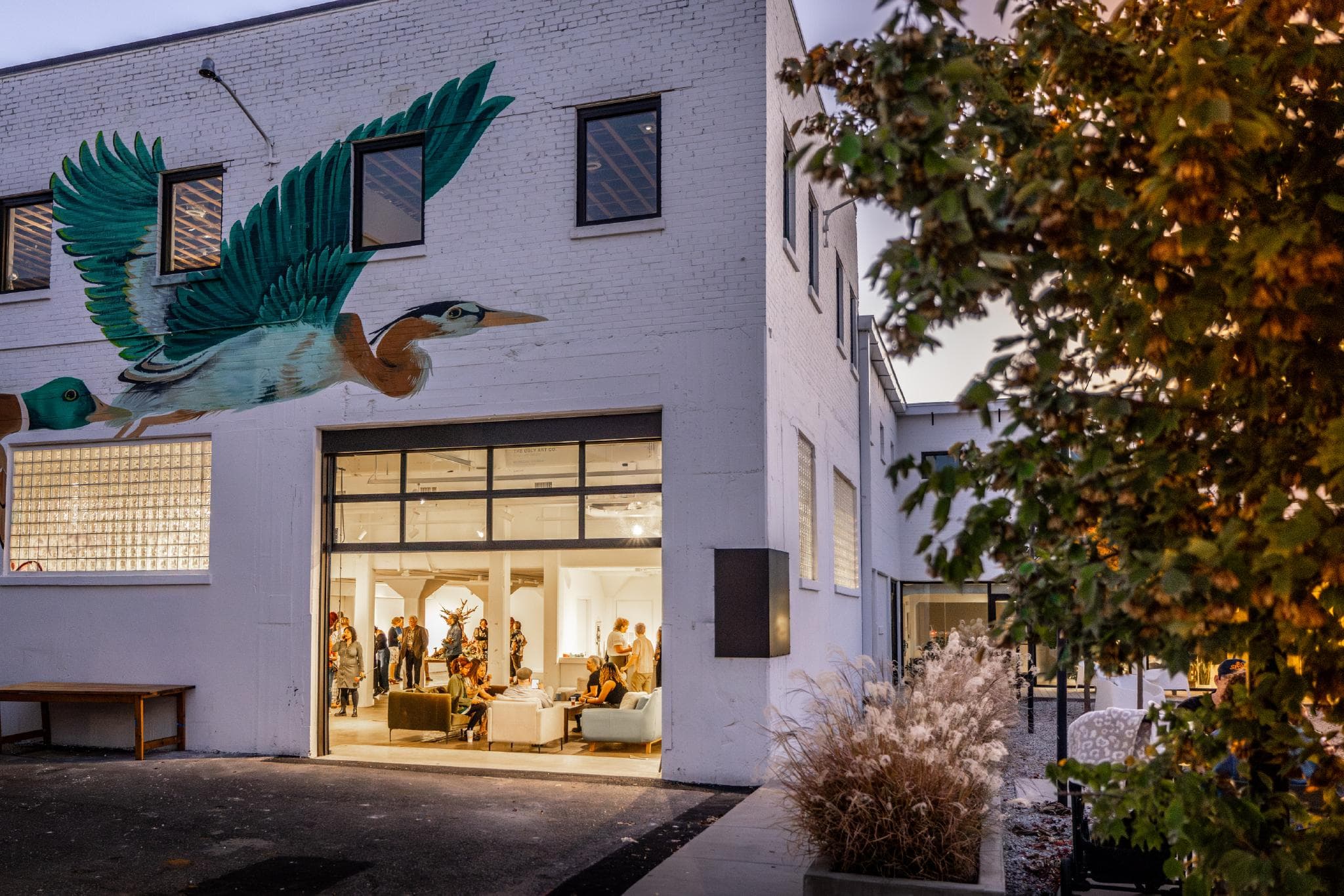 The exterior of Ugly Art Co., with a large crane mural over a large doorway. Inside, you can see people gathered for an event. 